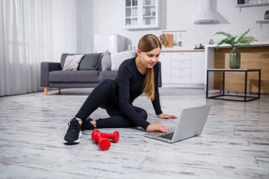 Siyah bluzlu ve taytlı genç bir bayan antrenör dizüstü bilgisayarla oturur ve karantina sırasında evde spor ve spor programı izler. Sağlıklı bir yaşam tarzı. Dambıllar yere düştü.