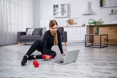 Siyah bluzlu ve taytlı genç bir bayan antrenör dizüstü bilgisayarla oturur ve karantina sırasında evde spor ve spor programı izler. Sağlıklı bir yaşam tarzı. Dambıllar yere düştü.