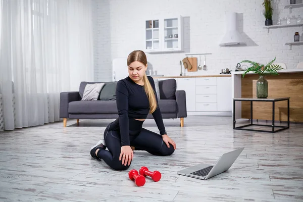Young sportive woman in fitness clothes in a modern home using online lessons from a fitness site in a laptop and doing workout at home. Sports at home during quarantine