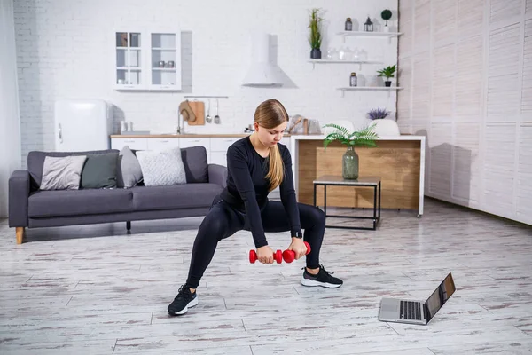 Young athletic woman in fitness clothes black top and leggings in an apartment using online workout from a fitness site on a laptop and doing sports at home. Fitness at home during quarantine
