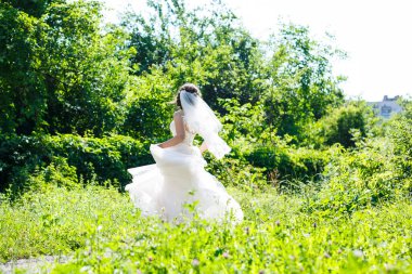 Uzun beyaz gelinlik içinde, yeşil bir parkta duvak takmış mutlu bir gelin..