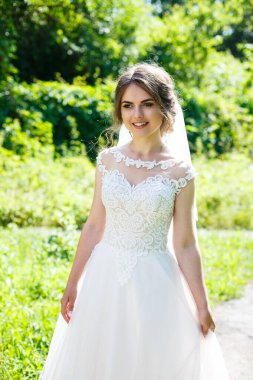 Uzun beyaz gelinlik içinde mutlu bir gelin ve doğa üzerine yeşil bir parkta duvak. Genç bir kızın düğün resmi, kadın makyajı ve saç stili. Evlilik kavramı