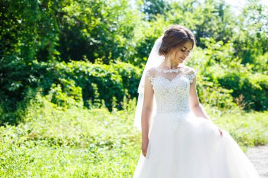 Uzun beyaz gelinlik içinde mutlu bir gelin ve doğa üzerine yeşil bir parkta duvak. Genç bir kızın düğün resmi, kadın makyajı ve saç stili. Evlilik kavramı