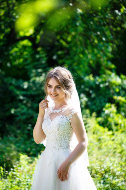 Uzun beyaz gelinlik içinde mutlu bir gelin ve doğa üzerine yeşil bir parkta duvak. Genç bir kızın düğün resmi, kadın makyajı ve saç stili. Evlilik kavramı