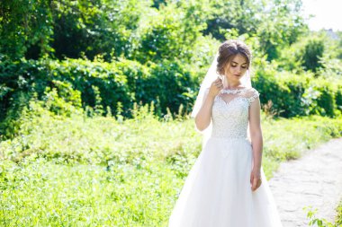 Uzun beyaz gelinlik içinde mutlu bir gelin ve doğa üzerine yeşil bir parkta duvak. Genç bir kızın düğün resmi, kadın makyajı ve saç stili. Evlilik kavramı