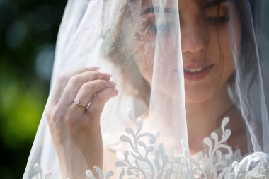 Uzun beyaz bir gelinlik içinde mutlu bir gelinin portresi ve yeşil bir parkta yüzü peçeyle. Genç bir kızın düğün resmi, kadın makyajı ve saç stili. Evlilik kavramı