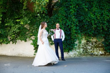 Ön planda beyaz elbiseli gelin ve arkasında pantolon askısı olan damat. Arkadaki duvarda yeşil yapraklar var.