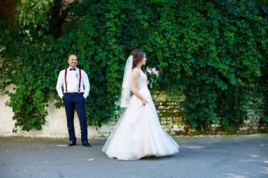 Ön planda beyaz elbiseli gelin ve arkasında pantolon askısı olan damat. Arkadaki duvarda yeşil yapraklar var.