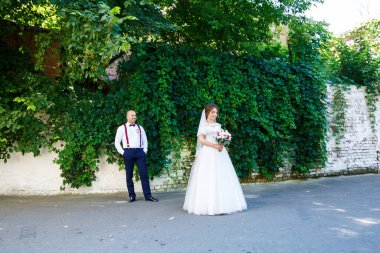 Ön planda beyaz elbiseli gelin ve arkasında pantolon askısı olan damat. Arkadaki duvarda yeşil yapraklar var.