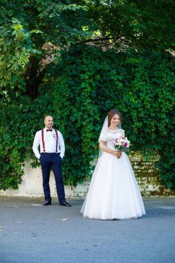 Ön planda beyaz elbiseli gelin ve arkasında pantolon askısı olan damat. Arkadaki duvarda yeşil yapraklar var.