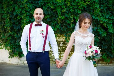 Pantolon askısı ve papyonlu damat el ele tutuşurken beyaz elbiseli güzel bir çift. Yeşil yapraklı bir duvarın arkasında. Mutlu bir çift. Evlilik kavramı.