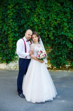 Damadın askısı ve papyonu varken beyaz elbiseli ve buketli neşeli çift. Yeşil yapraklı bir duvarın arkasında. Mutlu bir çift. Evlilik kavramı..