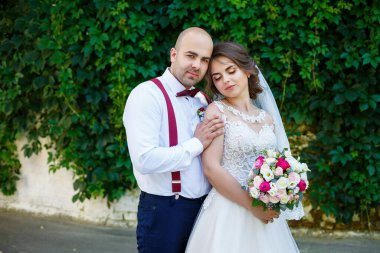 Damadın askısı ve papyonu varken beyaz elbiseli ve buketli neşeli çift. Yeşil yapraklı bir duvarın arkasında. Mutlu bir çift. Evlilik kavramı..