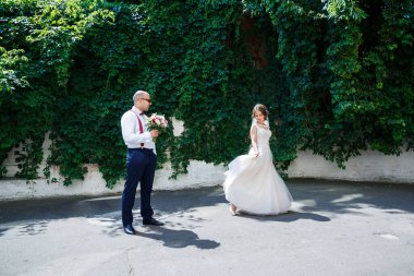 Beyaz gömlekli ve pantolonlu bir damat, beyaz elbiseli ve peçeli güzel bir gelin, düğün günü sokakta poz veriyor ve yürüyor.