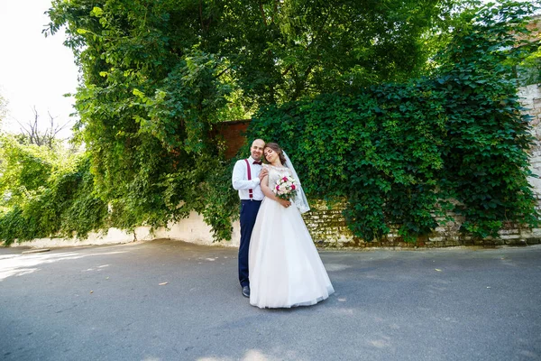 Damadın askısı ve papyonu varken beyaz elbiseli ve buketli neşeli çift. Yeşil yapraklı bir duvarın arkasında. Mutlu bir çift. Evlilik kavramı..