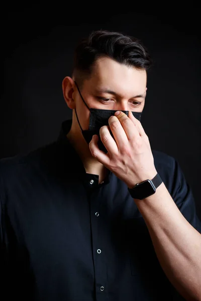 A young male doctor in a black surgical suit puts on a black mask to protect against the virus. Isolated on black background