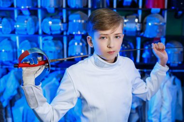 Elinde kılıç tutan, eskrim kıyafetli genç bir adam, mavi arka planda, neon ışıklı. Sporcu antrenman yapar. Spor, gençlik, sağlıklı yaşam tarzı.