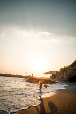 Siyah mayo ve gözlüklü şapkalı güzel bir kadın gün batımında kumsalda yürüyor. Deniz eğlencesi konsepti. Seçici odak