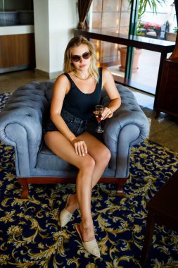 Güzel genç bir kadın, otel lobisindeki yumuşak bir koltukta elinde bir bardak şampanyayla oturuyor. O, güneş gözlüğü takıyor ve yüzünde bir gülümseme var. Tatil kavramı