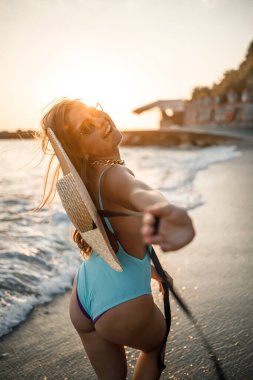 Gün batımında, mayolu ve gözlüklü şapkalı bir kadın kumsalda yürüyor. Deniz eğlencesi konsepti. Seçici odak