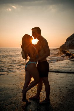 Deniz kenarında gün batımının arka planında birbirine aşık güzel bir çift. Genç bir kadın ve erkek gün batımında deniz kenarında sarılıyor.