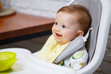 Dokuz aylık gülümseyen bir bebek, beyaz bir masada bir sandalyede oturur ve kaseden kaşıkla yemek yer. Anne bebeği kaşıkla besler. Arka plan bulanık. Çocuklar için sağlıklı yiyecekler. Çocuk maması..