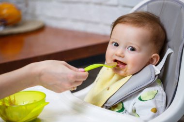 Dokuz aylık gülümseyen bir bebek, beyaz bir masada bir sandalyede oturur ve kaseden kaşıkla yemek yer. Anne bebeği kaşıkla besler. Arka plan bulanık. Çocuklar için sağlıklı yiyecekler. Çocuk maması..