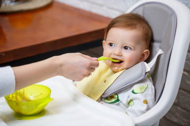 Dokuz aylık gülümseyen bir bebek, beyaz bir masada bir sandalyede oturur ve kaseden kaşıkla yemek yer. Anne bebeği kaşıkla besler. Arka plan bulanık. Çocuklar için sağlıklı yiyecekler. Çocuk maması..
