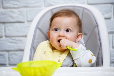 Dokuz aylık gülümseyen bir bebek, beyaz bir masada bir sandalyede oturur ve kaseden kaşıkla yemek yer. Anne bebeği kaşıkla besler. Arka plan bulanık. Çocuklar için sağlıklı yiyecekler. Çocuk maması..
