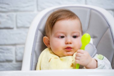 Dokuz aylık gülümseyen bir bebek, beyaz bir masada bir sandalyede oturur ve kaseden kaşıkla yemek yer. Anne bebeği kaşıkla besler. Arka plan bulanık. Çocuklar için sağlıklı yiyecekler. Çocuk maması..