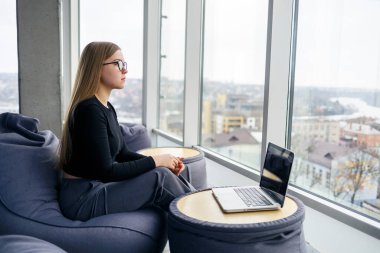 Güzel genç bir bayan yönetici, panoramik pencerenin yanındaki yumuşak bir puf üzerinde dizüstü bilgisayarla oturuyor. Kız işadamı yeni bir proje üzerinde çalışıyor.