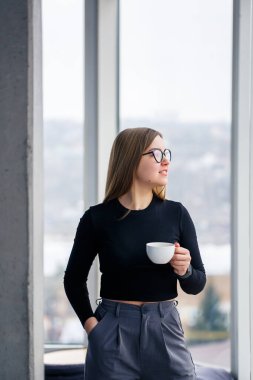 Profesyonel yetişkin bir iş kadını büyük cam pencerelerin önünde gömlek ve pantolonla kahve içiyor..