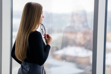 Kendi ofisinde büyük pencereli başarılı bir bayan yönetici duruyor ve şehre bakıyor. Büyük pencerelerin arkasında gözlüklü bir iş kadını.