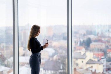 Kendi ofisinde büyük pencereli başarılı bir bayan yönetici duruyor ve şehre bakıyor. Büyük pencerelerin arkasında gözlüklü bir iş kadını.