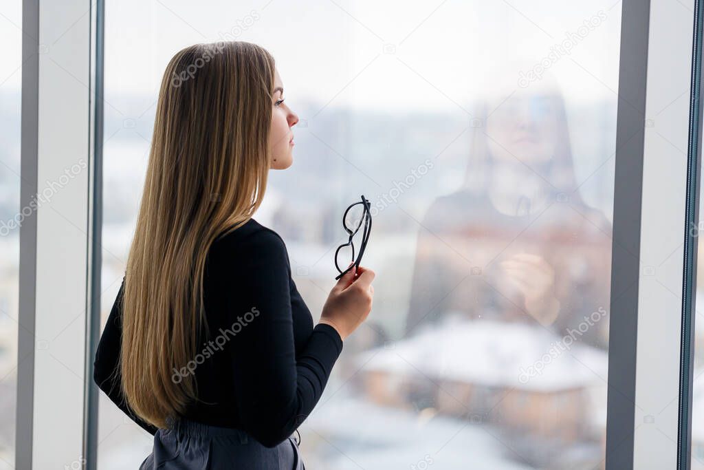 A successful female manager in her own office with large windows is standing and looking at the city. Business woman with glasses on the background of large windows