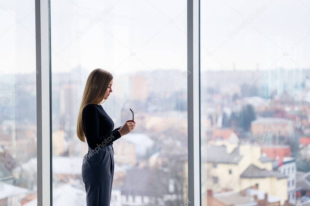 A successful female manager in her own office with large windows is standing and looking at the city. Business woman with glasses on the background of large windows