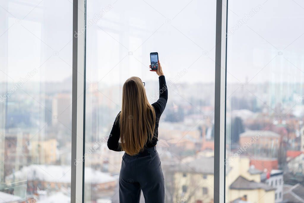 A successful female manager in her own office with large windows is standing and looking at the city. Business woman with glasses on the background of large windows
