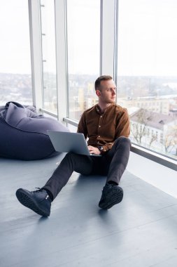Genç ve başarılı bir erkek işadamı büyük bir pencerenin yanında dizüstü bilgisayarla oturuyor. Uzak çalışma
