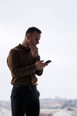 Genç ve başarılı bir erkek işadamı büyük bir pencerenin yanında gevşek bir şekilde duruyor ve şehre bakıyor..