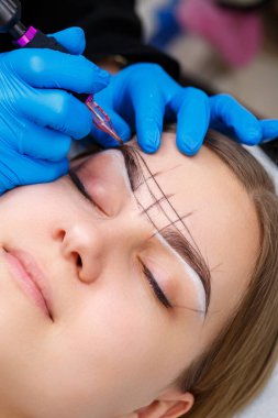 Güzellik uzmanı, kalıcı makyaj ve yakın çekim sırasında özel ekipman kullanarak bir kadının kaşlarına dövme yaptırıyor..