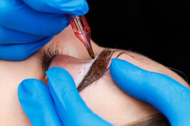 Güzellik uzmanı, kalıcı makyaj ve yakın çekim sırasında özel ekipman kullanarak bir kadının kaşlarına dövme yaptırıyor..