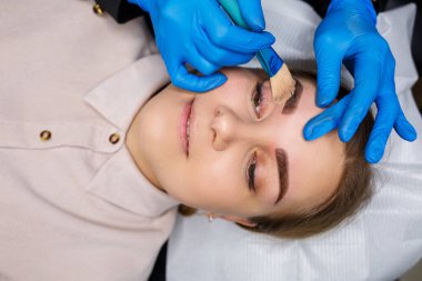 Kaşlarında kalıcı makyaj dövmesi olan bir kadın. Yakın plan güzellik uzmanı makyajı bir temele uygular. Profesyonel makyaj ve estetik cilt bakımı.