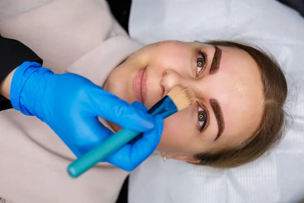 Woman with permanent makeup tattoo on her eyebrows. Close-up beautician makes makeup applies a foundation. Professional make-up and cosmetic skin care.