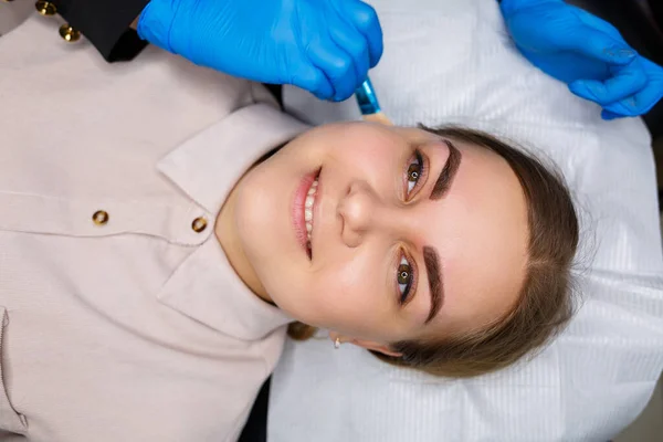 Woman with permanent makeup tattoo on her eyebrows. Close-up beautician makes makeup applies a foundation. Professional make-up and cosmetic skin care.