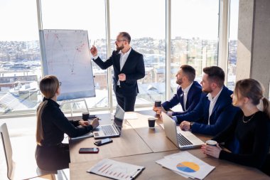 Confident businessman makes a presentation of a new project in the boardroom at a company meeting. Beautiful auditors talk with different partners about the business using a whiteboard and graphs. clipart