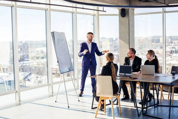 Confident businessman makes a presentation of a new project in the boardroom at a company meeting. Beautiful auditors talk with different partners about the business using a whiteboard and graphs.