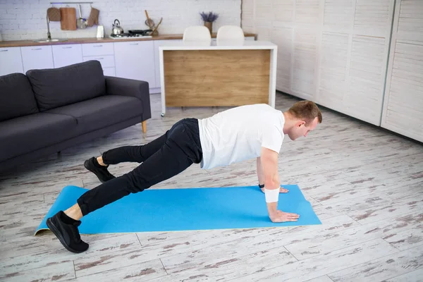 Handsome muscular man in a t-shirt doing functional plank exercises on the floor at home. Fitness at home. Healthy lifestyle.