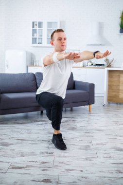 Tişörtlü yakışıklı adam evinde çömelmiş vaziyette oturuyor. Evde fitness var. Sağlıklı yaşam tarzı