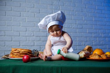 Önlük giymiş güzel bir çocuk, çocuk pişirme kostümü. Masada krep ve taze hamur işleri var.