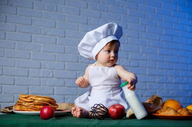 Önlük giymiş güzel bir çocuk, çocuk pişirme kostümü. Masada krep ve taze hamur işleri var.